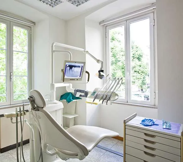 A quiet, sunlit dental treatment room with a modern chair and monitor, overlooking green foliage through a bright window.
