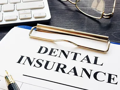 A clipboard reading DENTAL INSURANCE on an office desk with a fountain pen, glasses, and a keyboard.