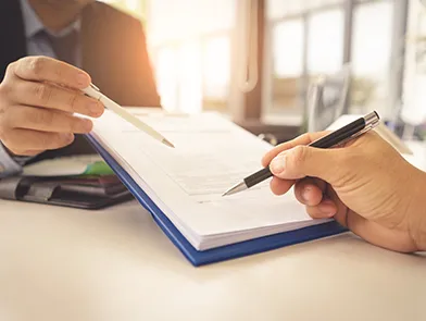 Close-up of hands signing a document on a blue clipboard in a bright office.
